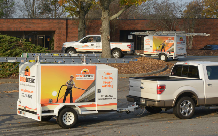 Two Outback GutterVac-branded pickup trucks are parked with their equipment trailers in front of a leaf-covered office park.