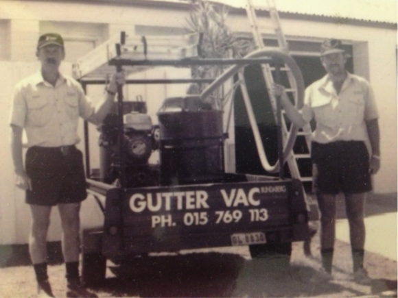 A sun-faded photograph of two GutterVac technicians standing next to a trailer with an industrial vacuum.