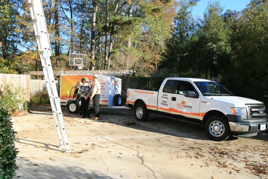 An Outback GutterVac technician speaks with a client in a leaf-covered driveway. A branded pickup truck and ladder are visible in the foreground.
