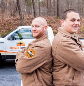 Two Outback GutterVac technicians stand smiling back-to-back in front of their branded pickup truck and a leafy forest.
