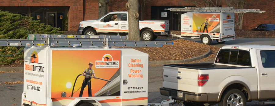 Two branded pickup trucks with equipment trailers and ladders are parked at a leaf-covered office park.