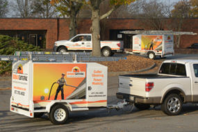 Two Outback GutterVac pickup trucks with equipment trailers sit parked outside a commercial building, with leaves littering the ground around them.
