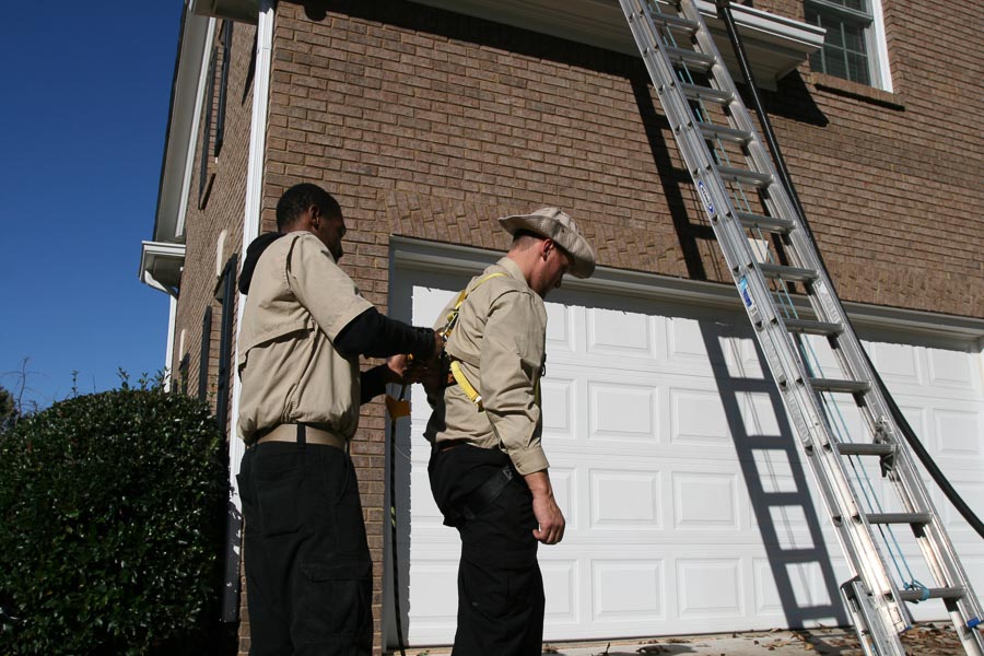 A technician hooks up safety equipment on another technician in front of a client’s home, with a ladder propped up against the garage in the background.