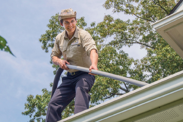 A smiling franchise owner cleans gutters with a vacuum on top of a client’s roof.