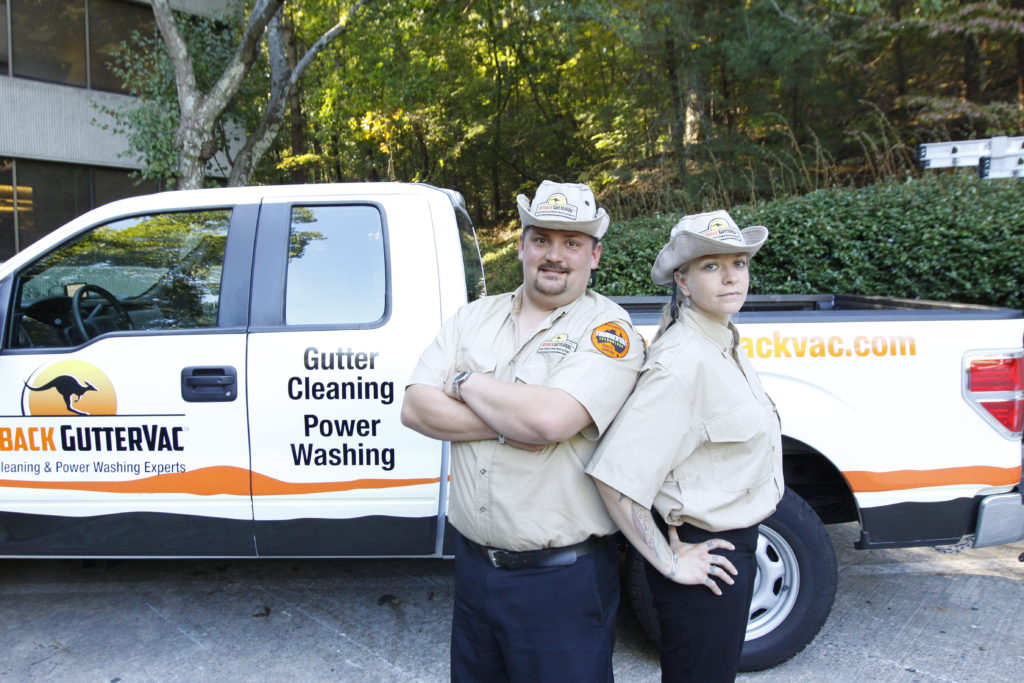 Two franchisees, one male and one female, stand in front of an Outback GutterVac-branded pickup truck, with “Gutter Cleaning Power Washing” printed on the side panel.