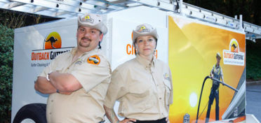 Two Outback GutterVac technicians pose in front of a branded equipment trailer.